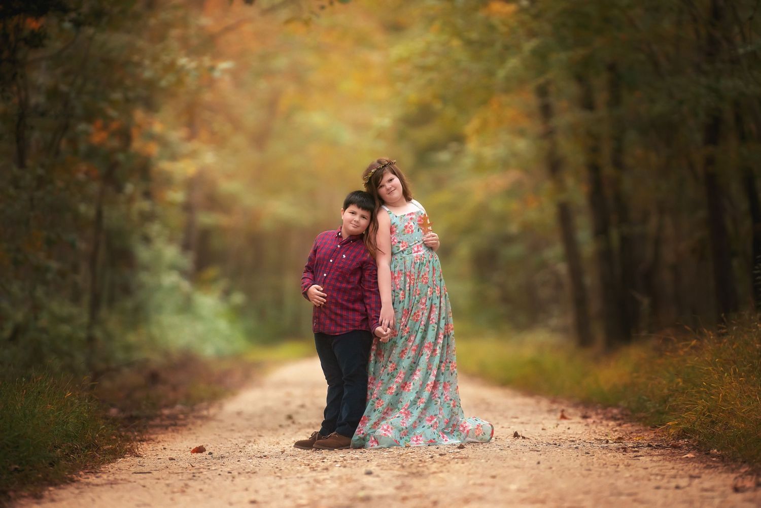 Pregnant woman in a floral maxi dress holds hands with a boy in a maroon shirt on a gravel path through autumn woods.