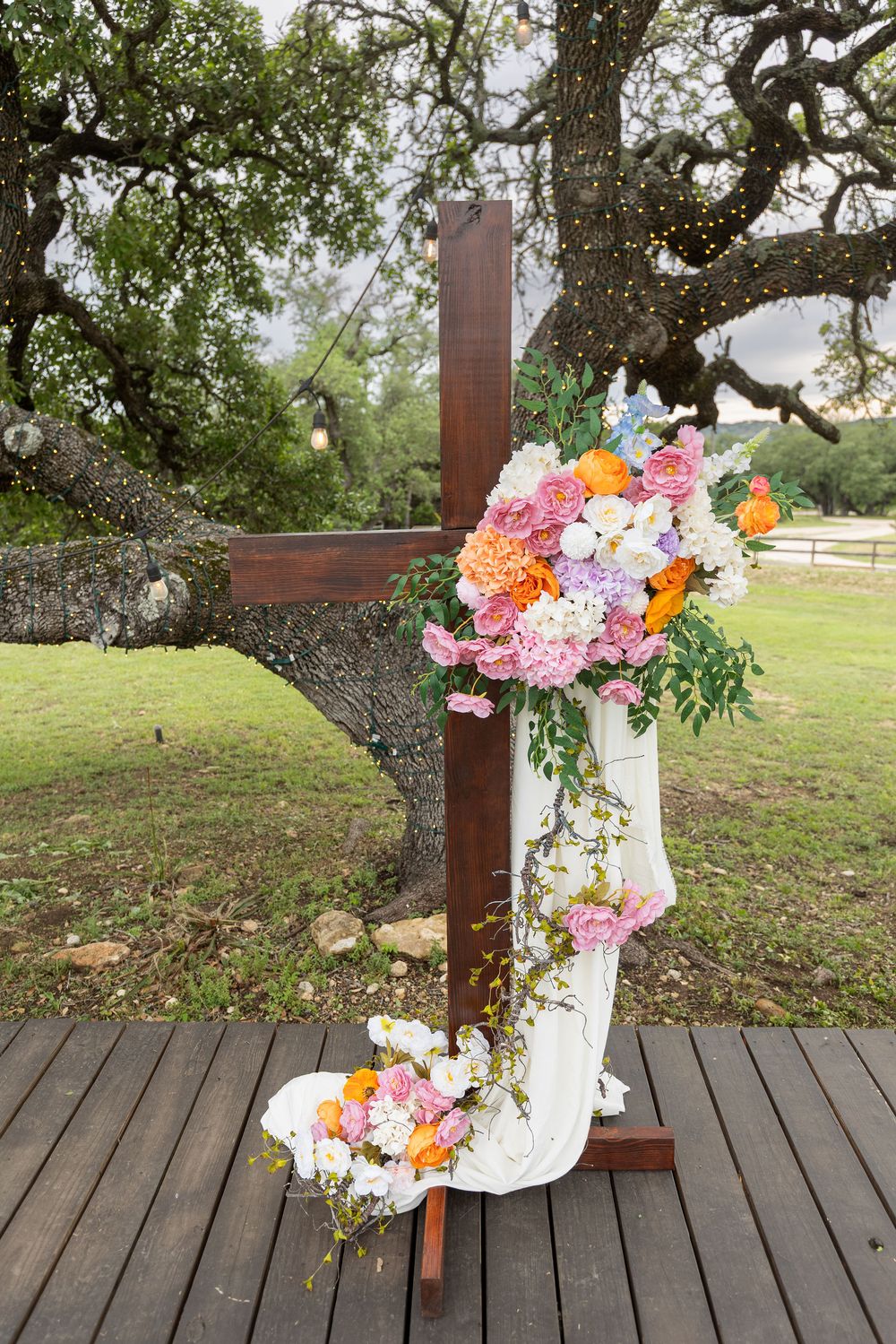 Wooden cross draped in white fabric with pink, orange and white flowers at an outdoor wedding ceremony beneath a string-lit oak tree.