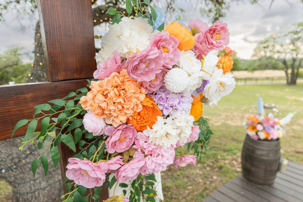 Lush wedding floral arrangement of pink, orange, white and purple blooms mounted on a wooden arch beside a rustic barrel outdoors.