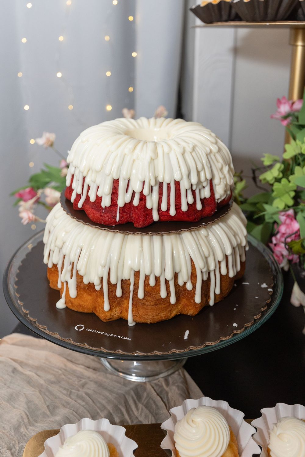Two-tiered bundt cake with red velvet top and golden bottom layer, covered in white icing drizzle, on a glass cake stand near pink flowers.