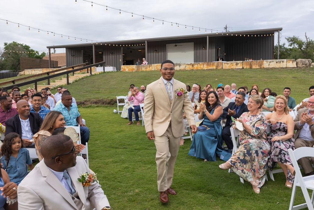 Smiling groom in a tan suit walks down the grass aisle past seated guests at an outdoor wedding ceremony with a rustic pavilion behind.