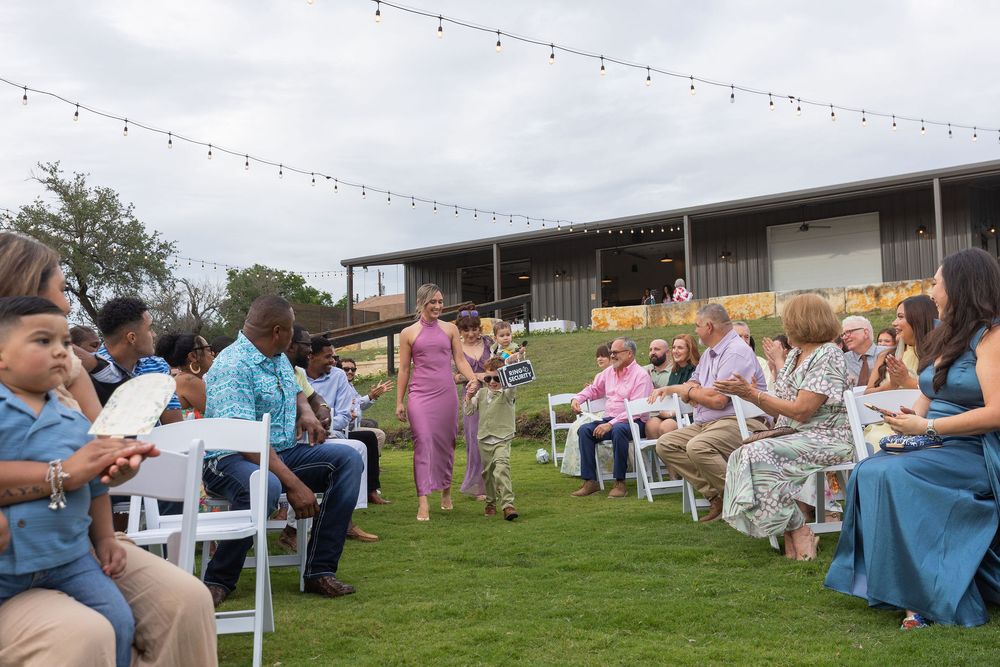 Guests seated on white chairs watch two women in dresses walk down the grass aisle at an outdoor wedding ceremony under string lights.