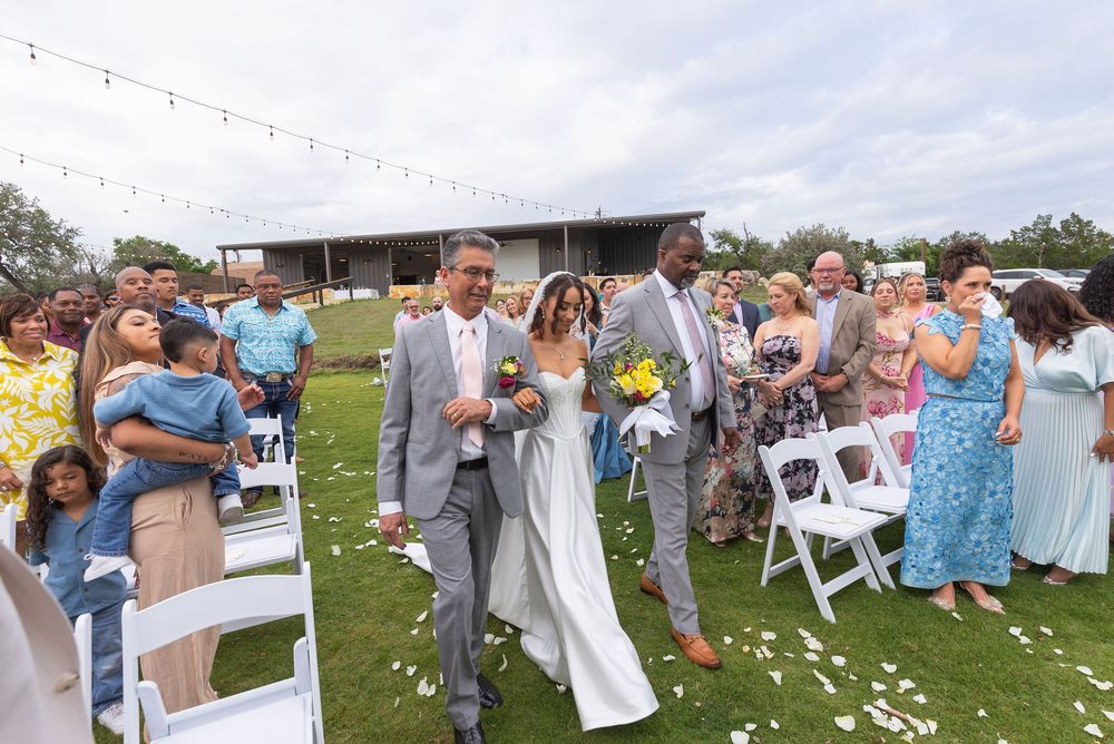Bride in a white gown walks down a grass aisle between two men during an outdoor wedding ceremony as smiling guests stand by white chairs.