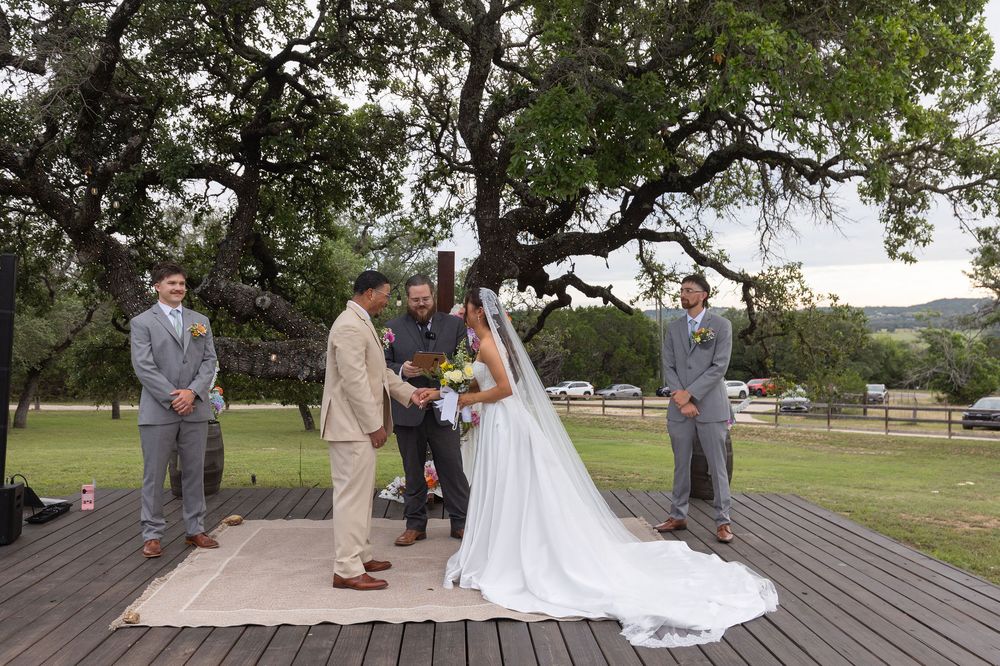 Bride in a white gown and veil exchanges vows with a groom on an outdoor wooden deck while groomsmen and an officiant stand under oak trees.