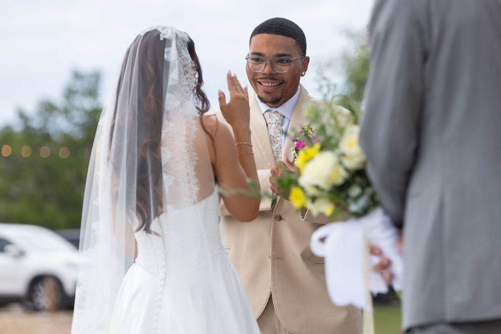 Smiling groom in a tan suit raises his hand as a bride in a long lace veil and white gown faces him during an outdoor wedding ceremony.