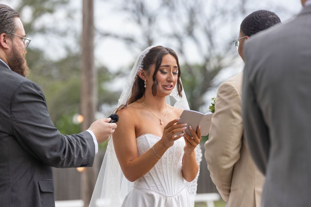 Emotional bride in a white strapless wedding gown and veil reads her vows during an outdoor ceremony as an officiant holds a microphone.