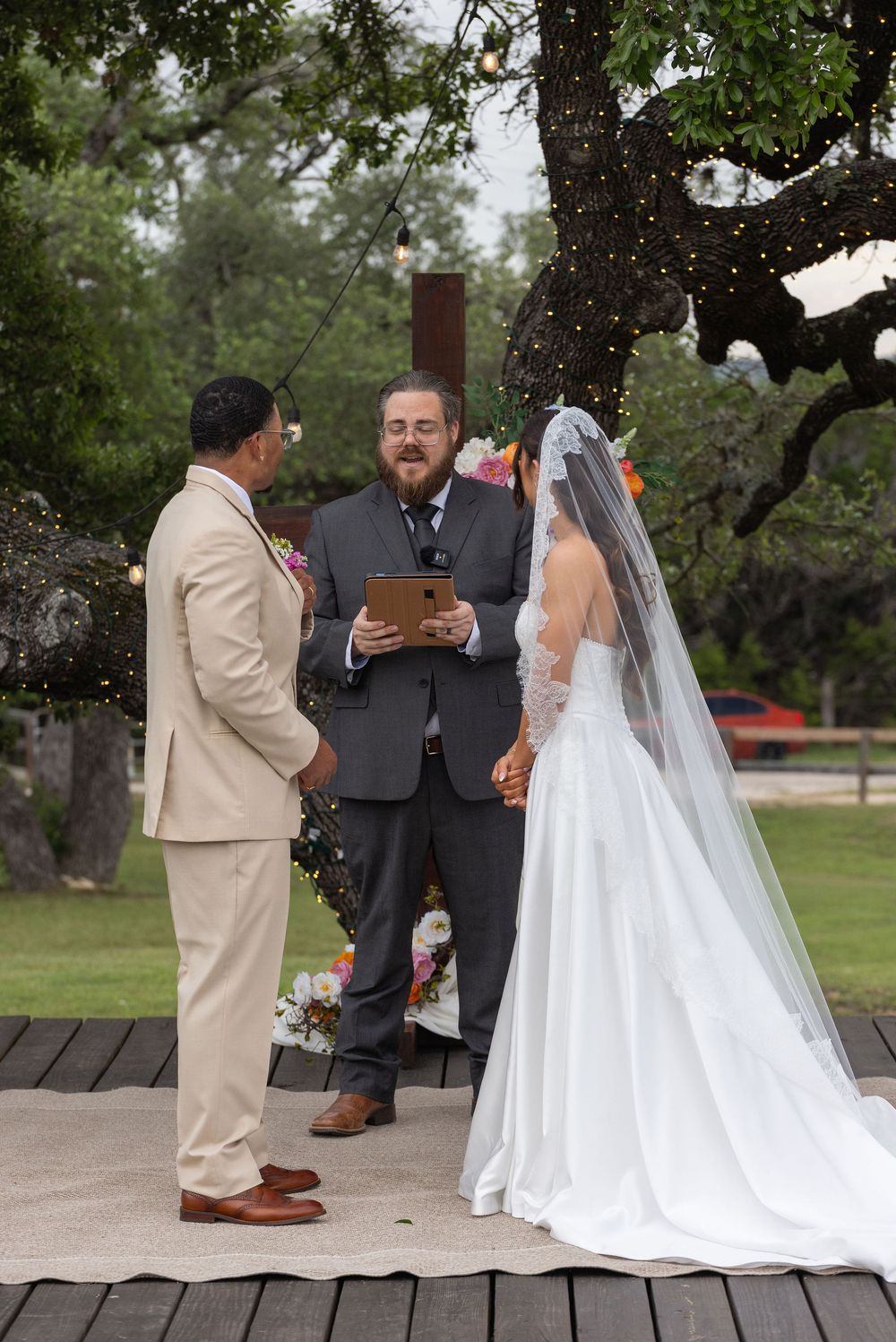 Bride in a long veil and white gown and groom in a tan suit exchange vows with an officiant outdoors under string lights and oak trees.