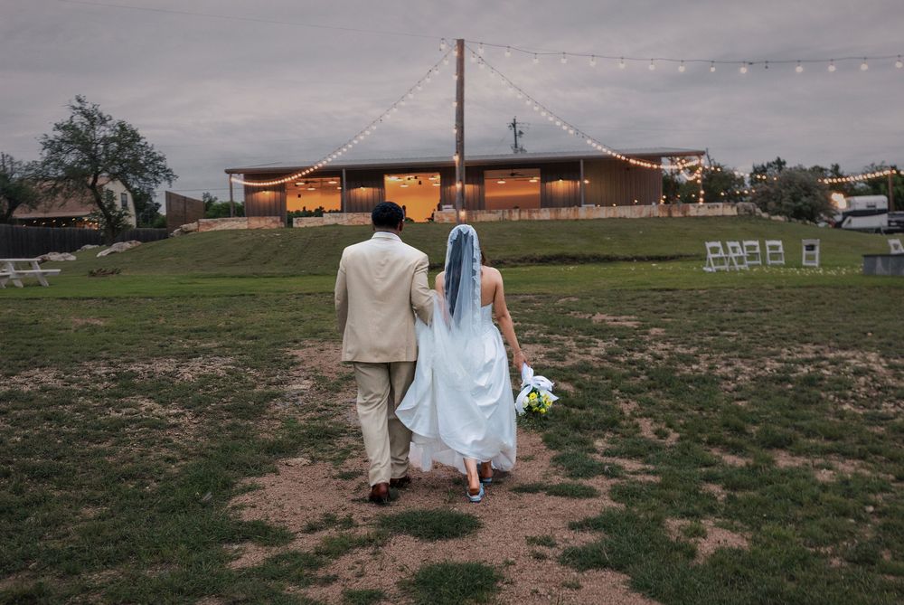 Bride in a long veil and groom in a tan suit walk hand in hand across a grassy field toward a rustic venue at dusk.