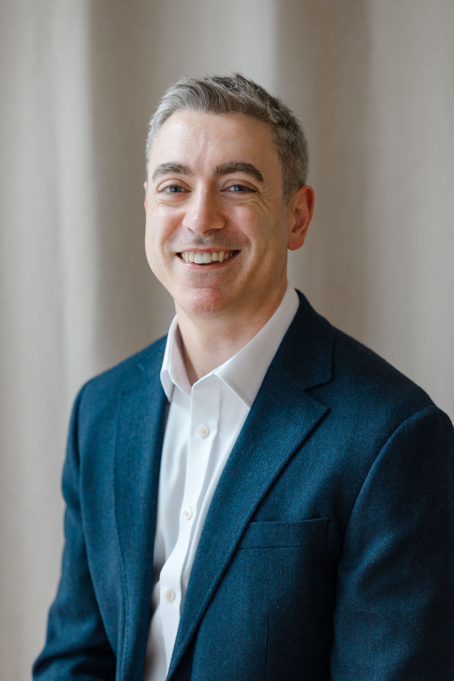 Smiling middle-aged man with gray hair wearing a teal blazer and white shirt poses for a professional headshot.
