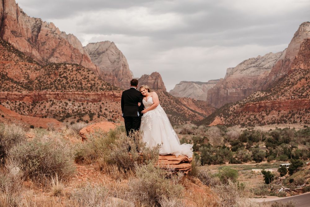 Bride and groom stand on red rock cliff overlooking stunning Zion National Park canyon landscape on their wedding day.