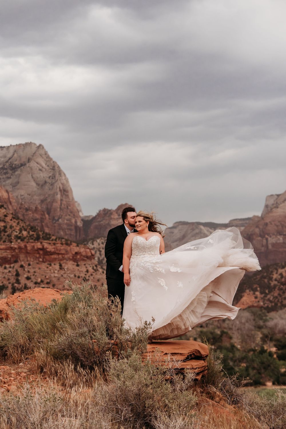 Bride and groom embrace on red rock cliffs at Zion National Park, wedding dress flowing in the wind under dramatic cloudy skies.