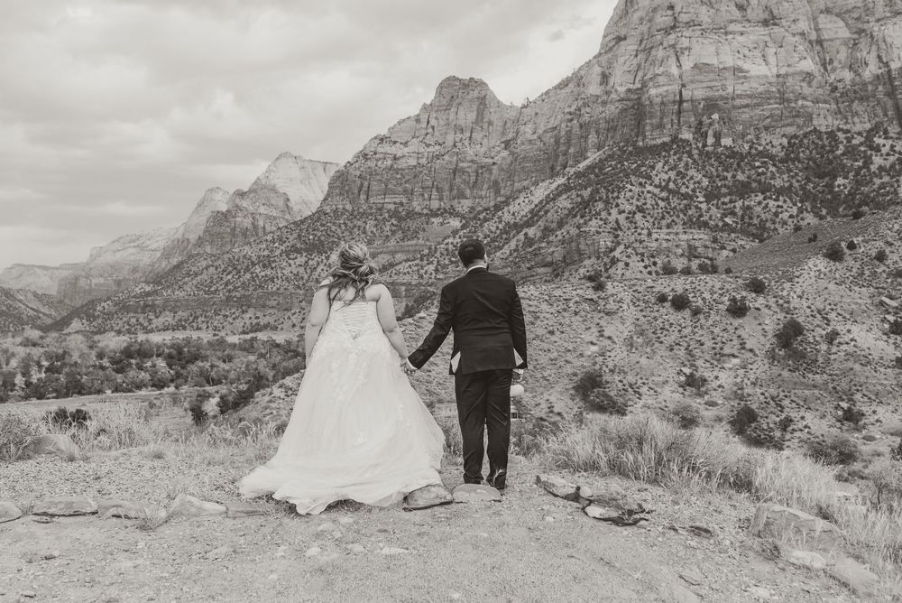 Black and white photo of bride and groom holding hands overlooking stunning Zion National Park canyon landscape.