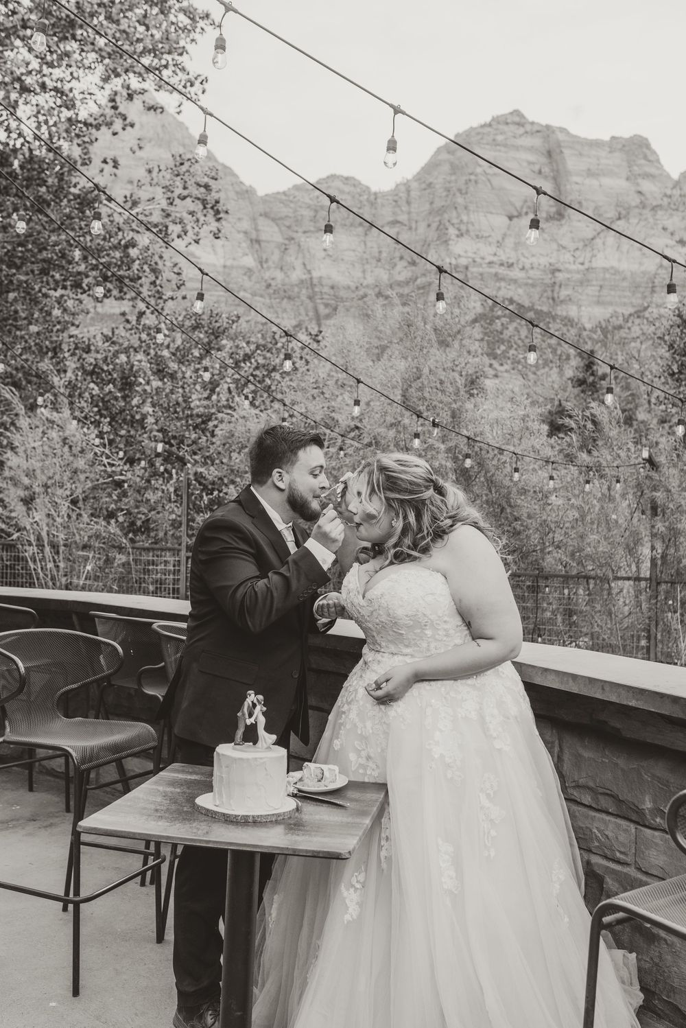 Black and white photo of bride and groom sharing wedding cake on mountain view terrace with string lights.