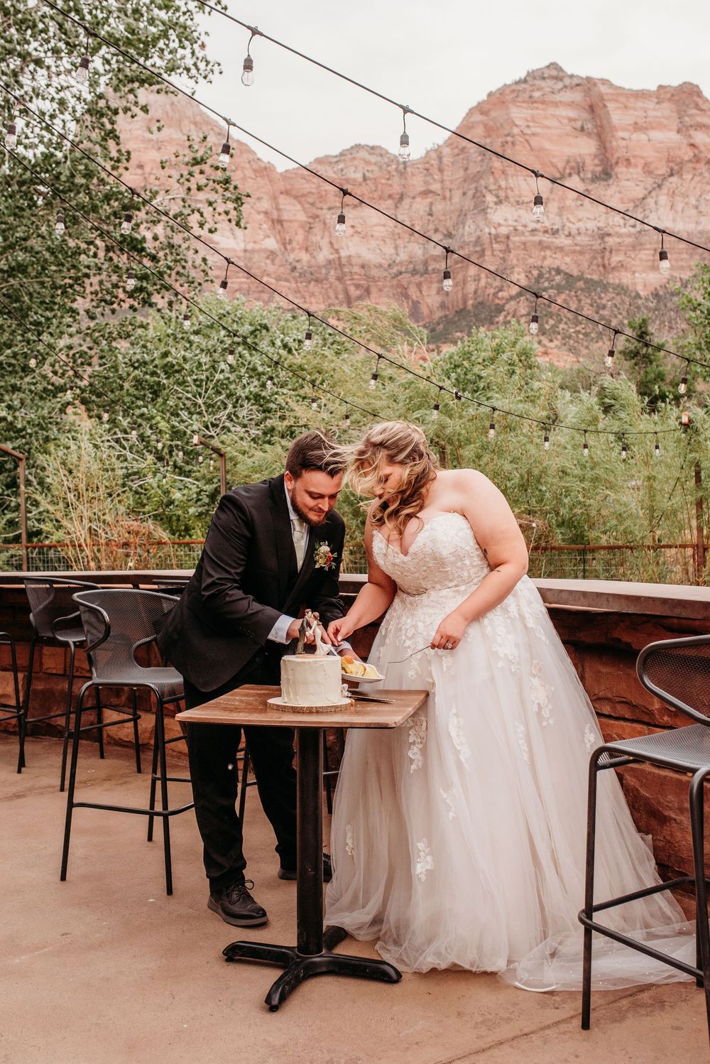 Bride and groom cutting wedding cake on outdoor patio with stunning red rock Zion National Park mountains backdrop.
