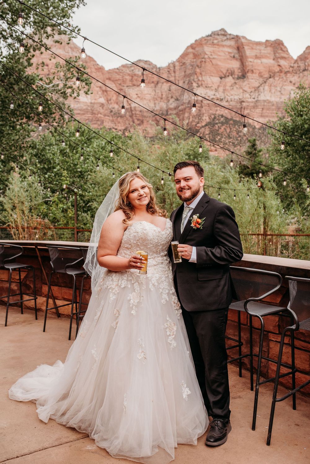 Bride and groom posing on a terrace with red rock Zion National Park mountains backdrop and string lights.