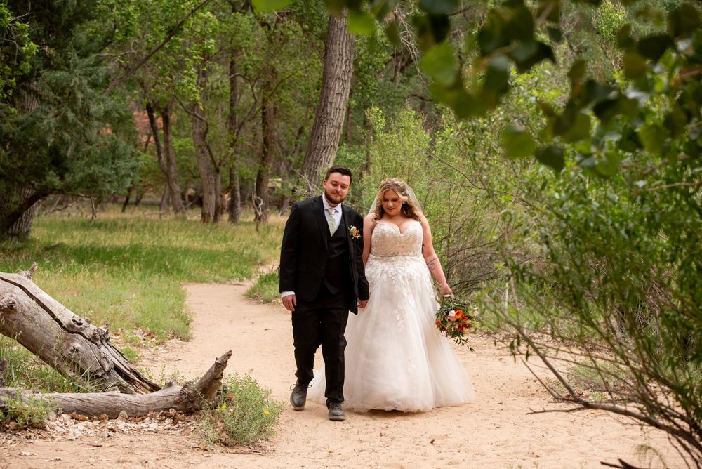 Bride and groom walking hand in hand on a sandy path surrounded by lush green trees during an outdoor wedding ceremony.