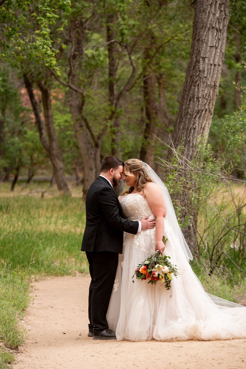 Bride and groom share romantic moment on forest path, bride holding vibrant bouquet in elegant lace ball gown.