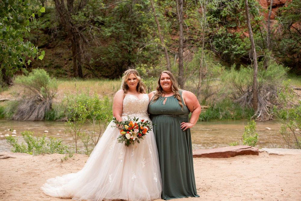 Bride in white gown holding bouquet stands with bridesmaid in green dress at outdoor Zion National Park wedding.