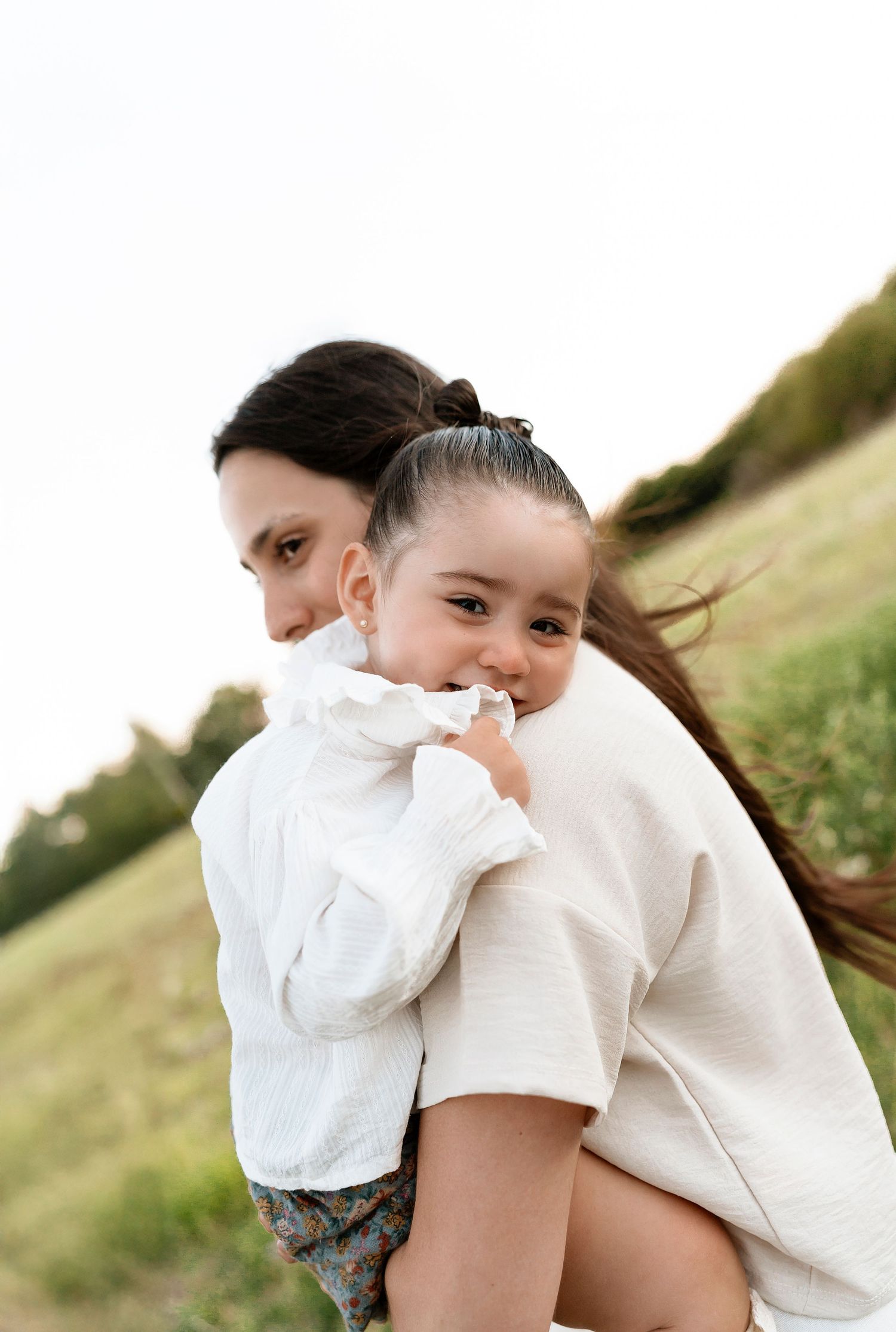 A woman hugging a young girl in a white dress outdoors in a green field.
