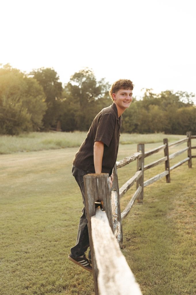 A senior boy leans on a rustic wooden fence in a sunny rural field with trees in the background