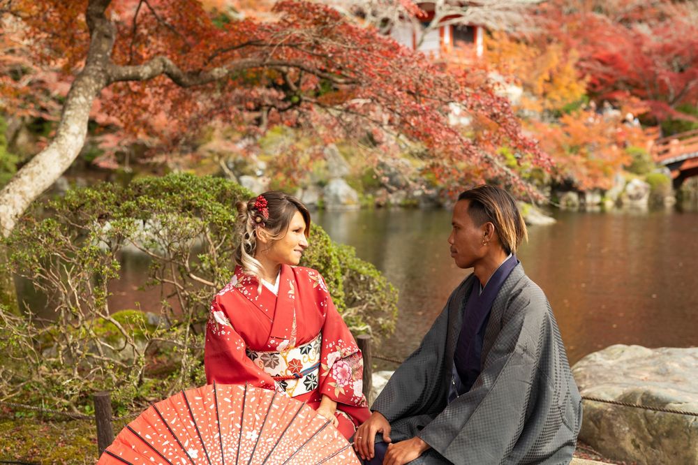 Couple in traditional Japanese kimono sitting beside a calm pond with vibrant red autumn maple leaves and a red parasol in the foreground.