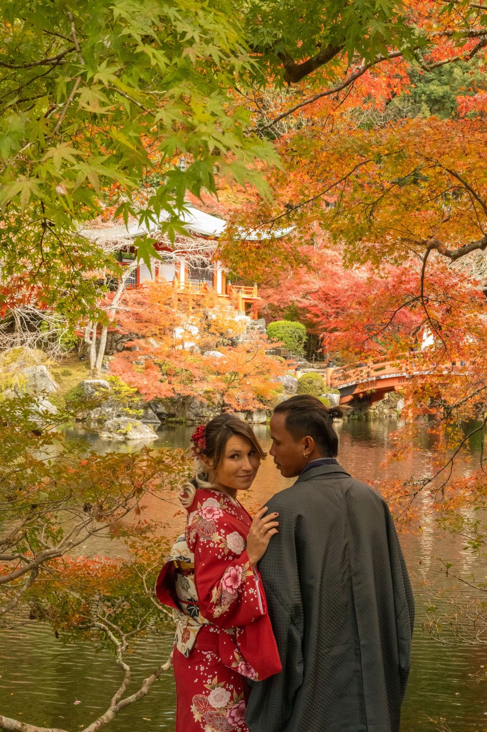 Couple in traditional Japanese kimono embracing beside a tranquil pond surrounded by vibrant red autumn maple leaves and a pavilion.