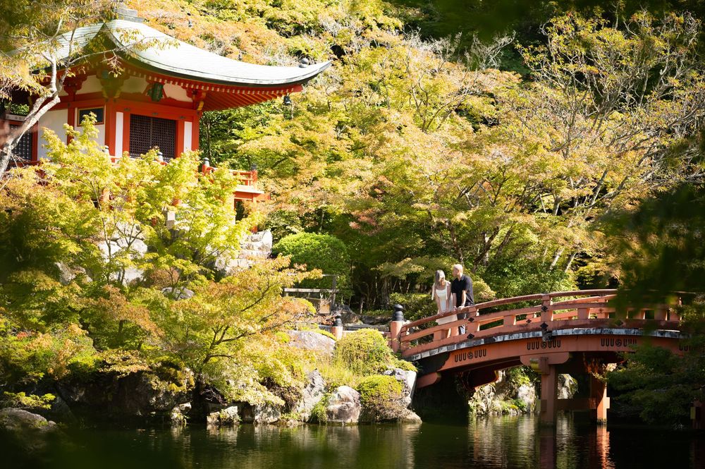 Two visitors stand on a red arched wooden bridge beside a pond, with a red-roofed Japanese temple surrounded by lush green autumn trees.