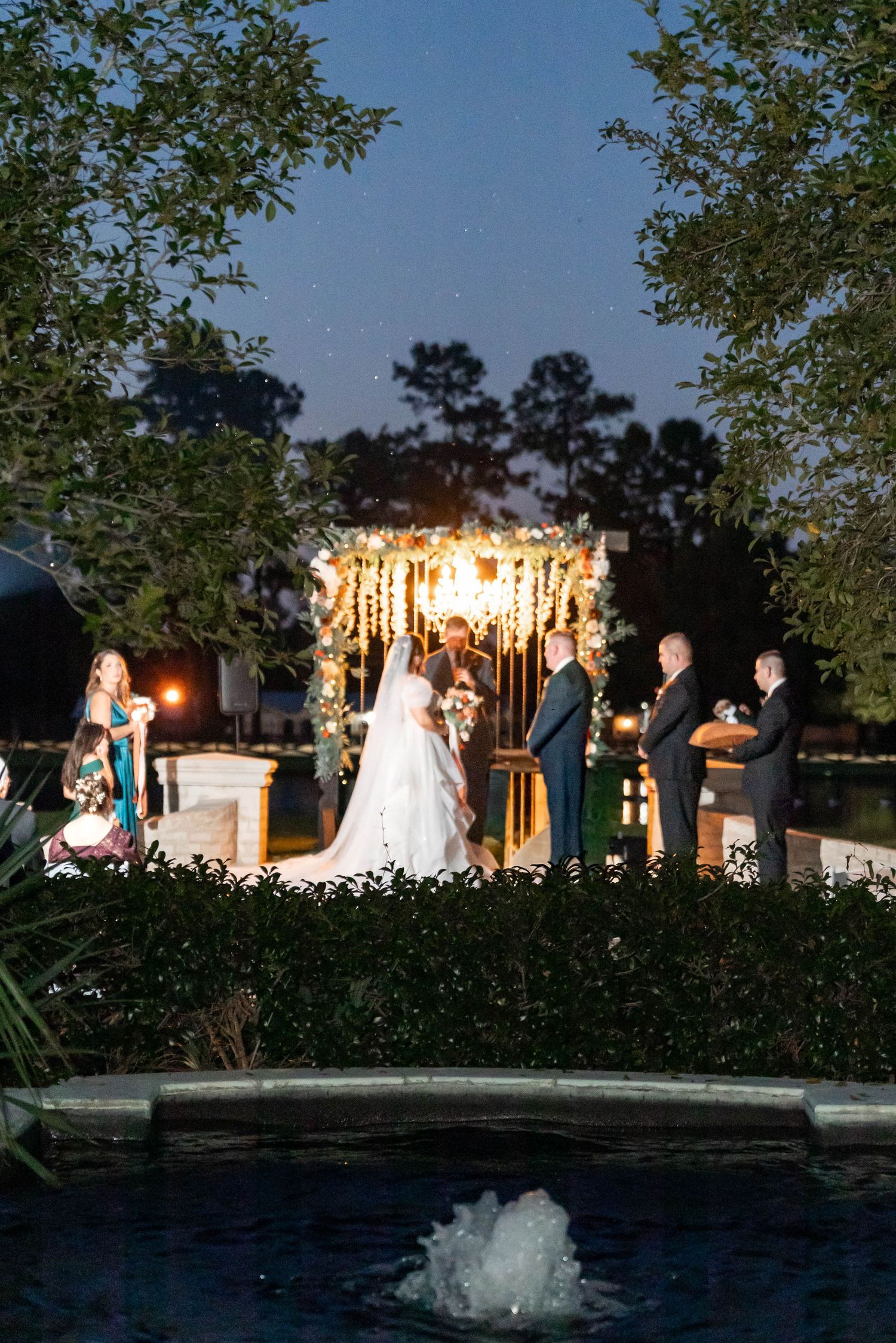 Bride and groom exchange vows under a lit floral arch at an outdoor evening wedding ceremony beside a fountain pool, with guests looking on.