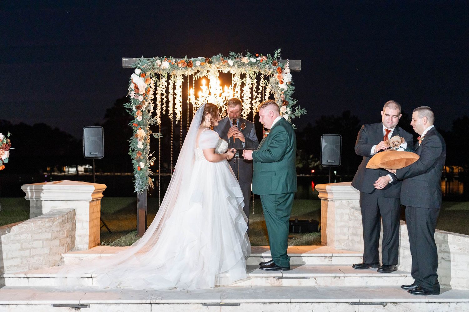 Bride in a flowing white gown and groom in a green suit exchange vows under a floral wedding arch at an outdoor evening ceremony.