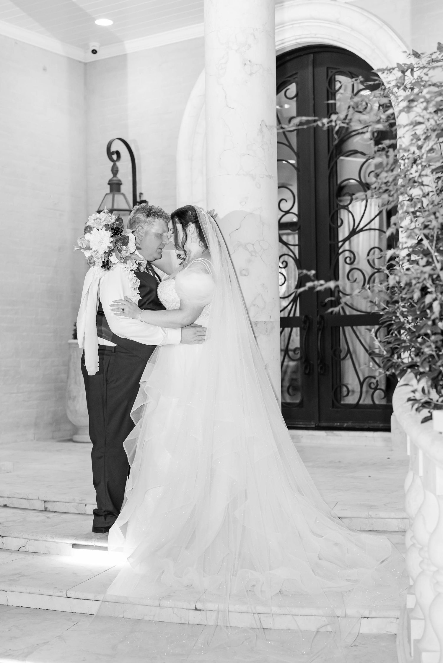 Black and white photo of a bride and groom embracing on stone steps beside a column and ornate wrought iron double doors.