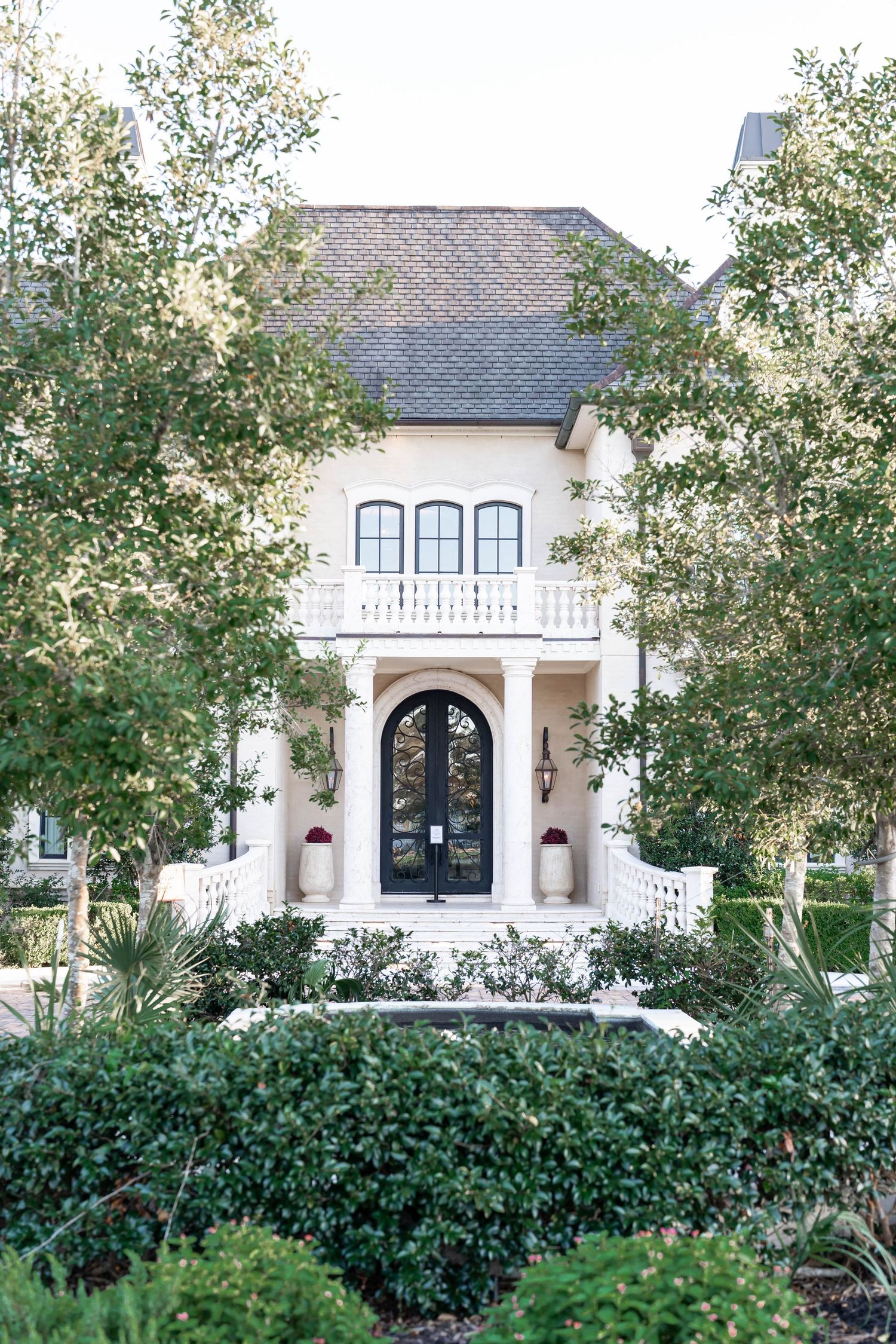 Elegant cream luxury home with arched black double doors, columned entry, white balustrade balcony, slate roof and lush landscaped greenery.