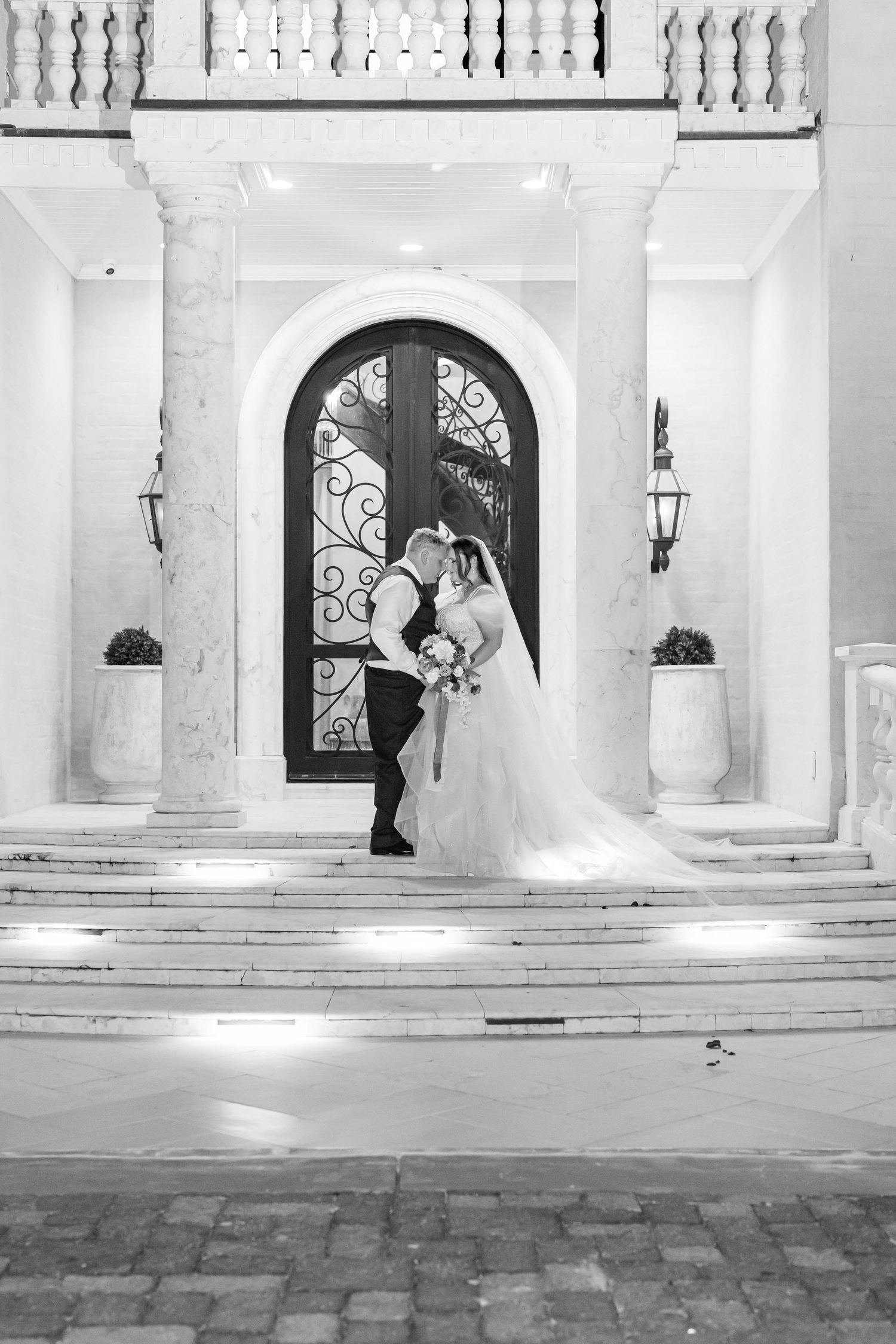 Black and white photo of a bride and groom embracing on marble steps before an ornate wrought iron arched door at an elegant wedding venue.