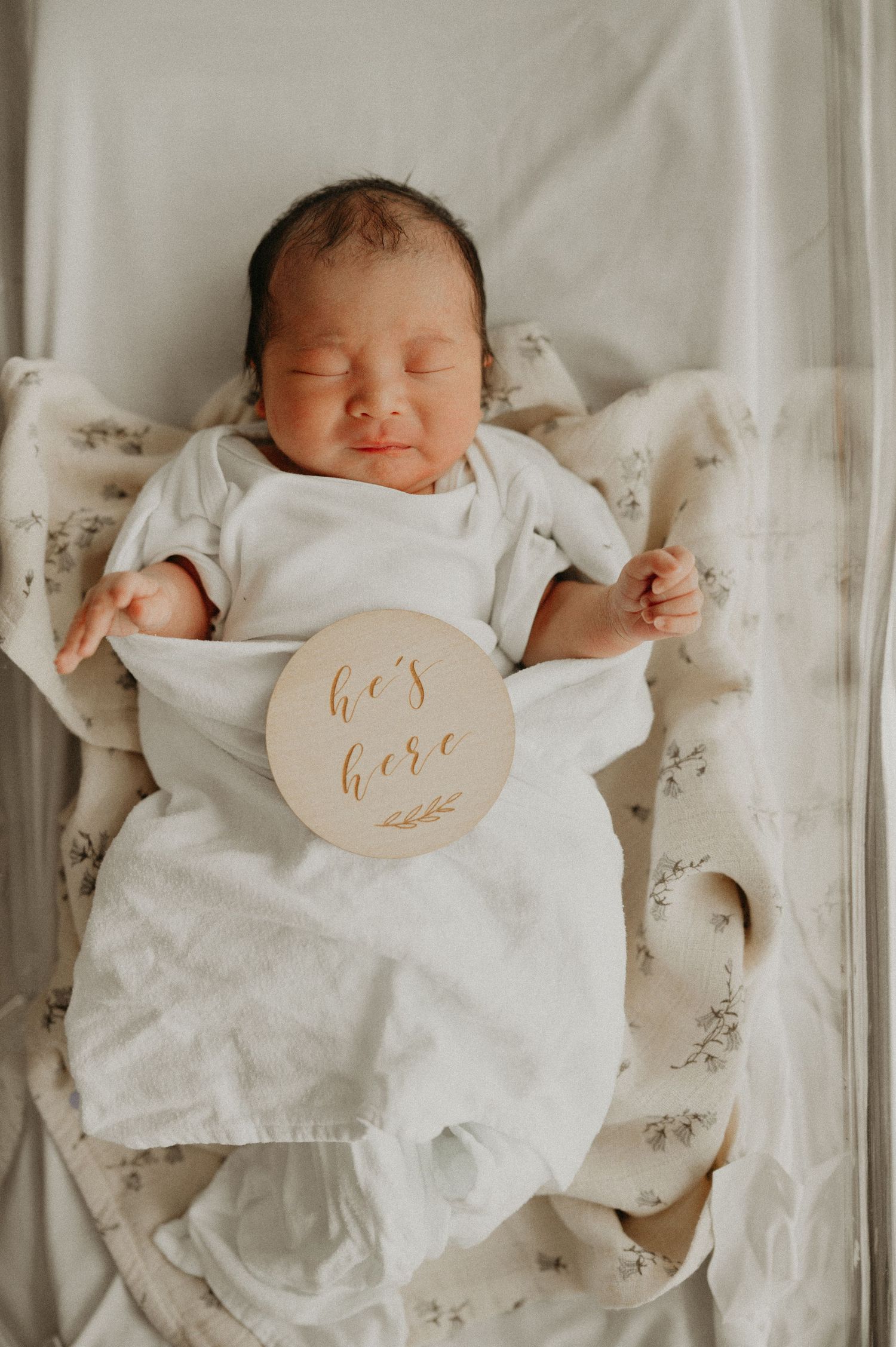 Sleeping newborn baby in a white swaddle on a floral blanket holding a wooden "he's here" birth announcement sign.
