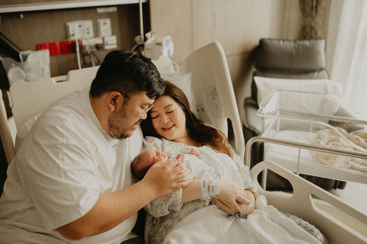New parents smiling down at their sleeping newborn baby while sitting together on a hospital bed in a bright maternity room.