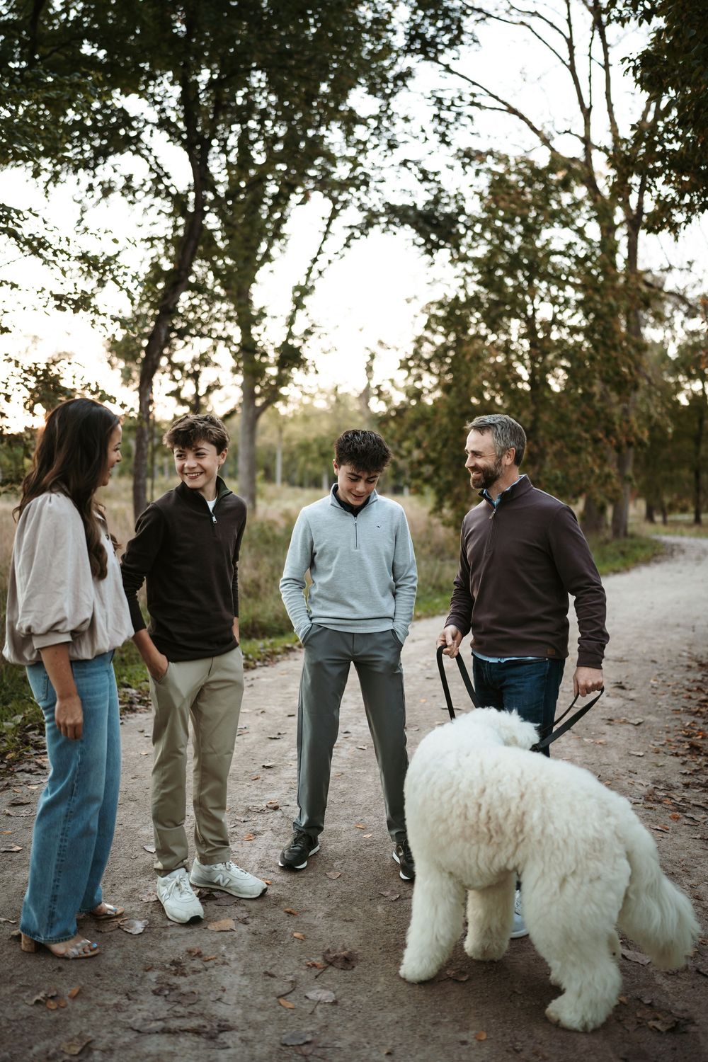 A family of four stands chatting on a  dirt path at sunset with their fluffy white dog on a leash a family fall session In Oakbrook