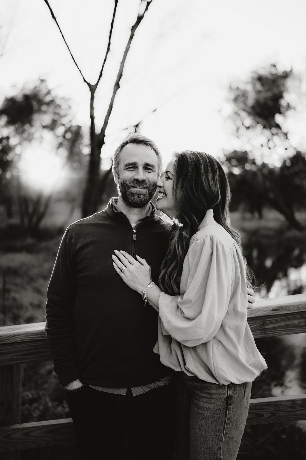 Black and white photo of a smiling couple embracing on a wooden bridge with warm sunlight glowing through trees behind them.