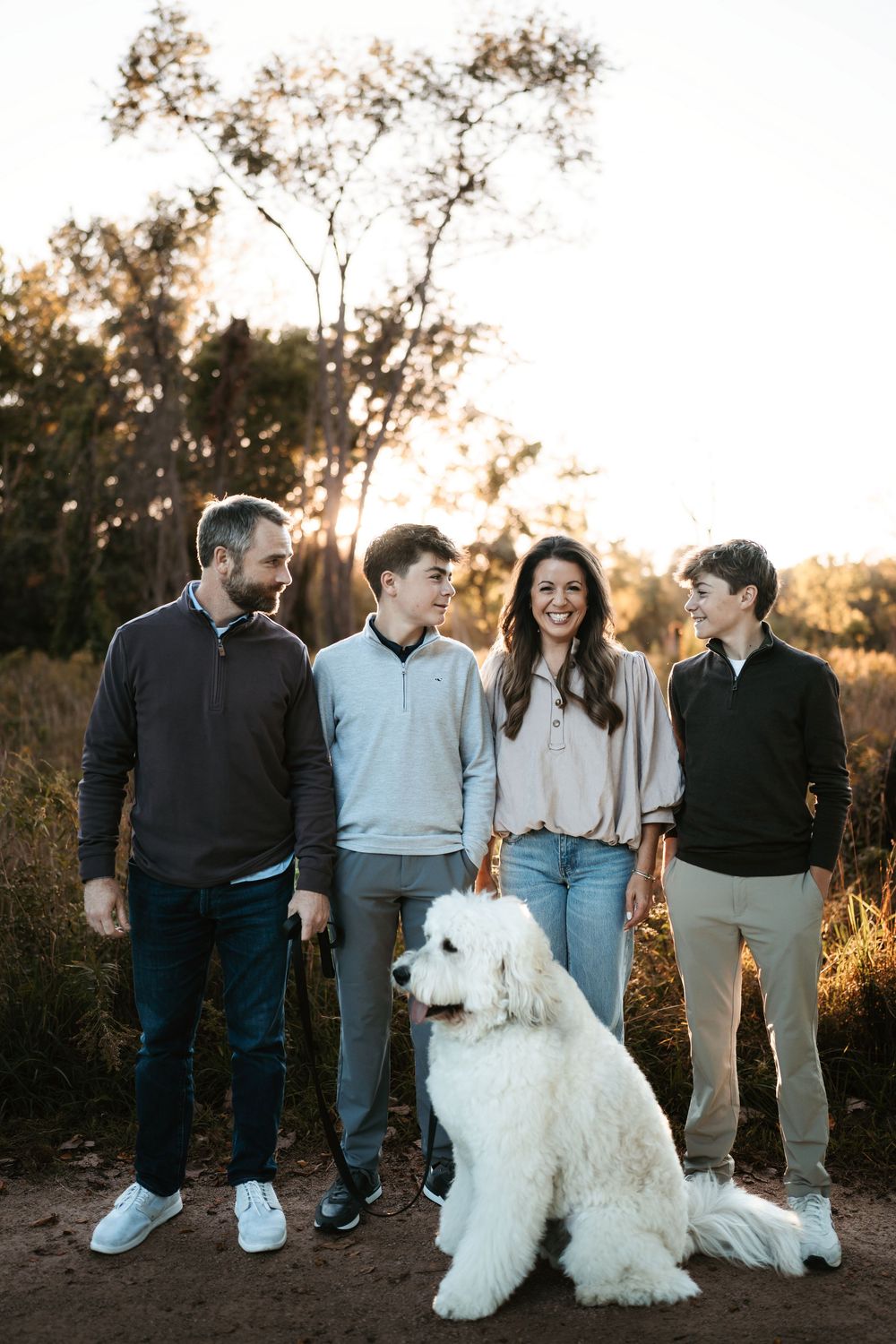 A family of four stands outdoors at golden hour with a large fluffy white dog sitting in front of them on a dirt path.