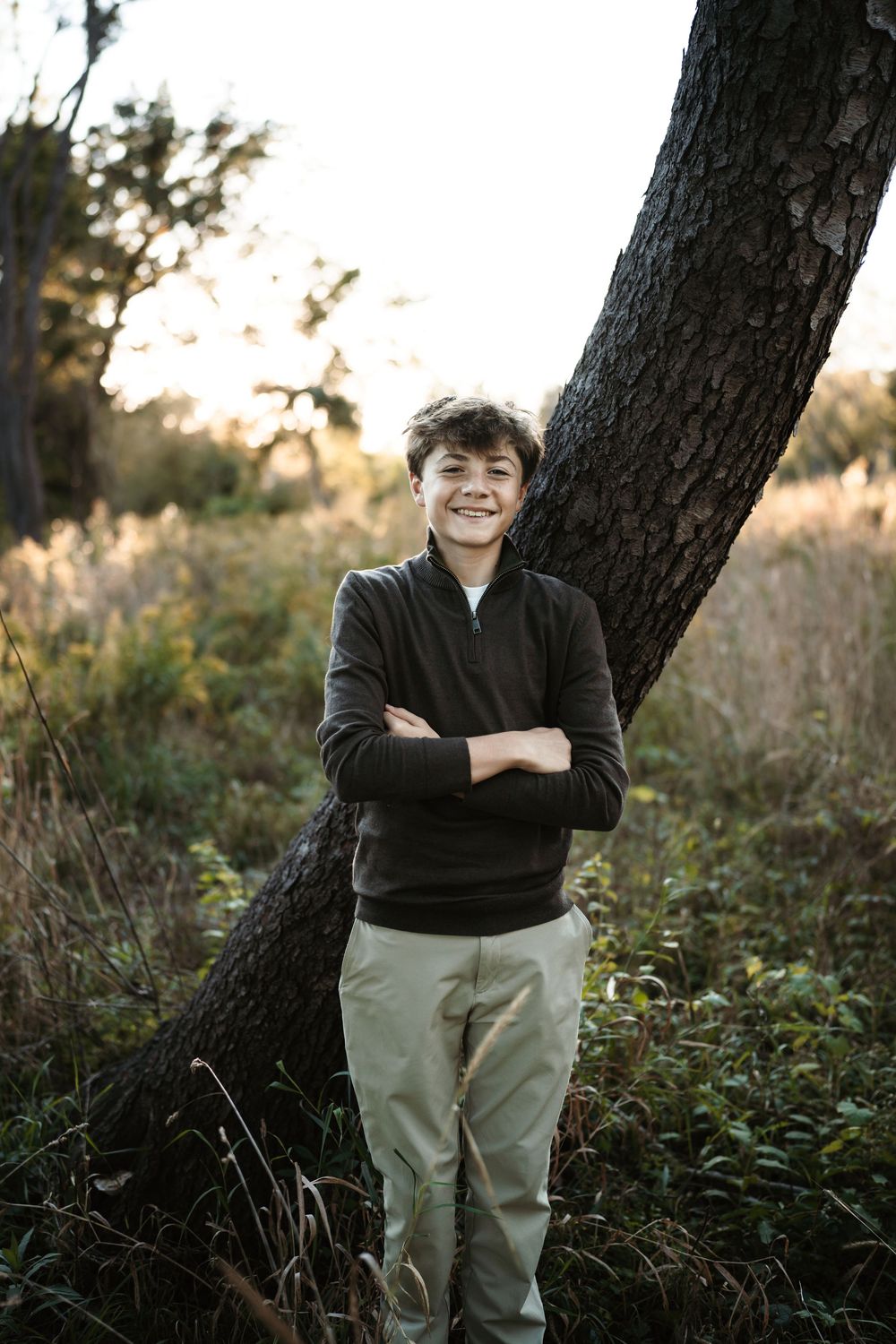 Smiling teenage boy in a dark zip-up pullover and khaki pants leans against a slanted tree trunk in a golden-lit grassy field at sunset.