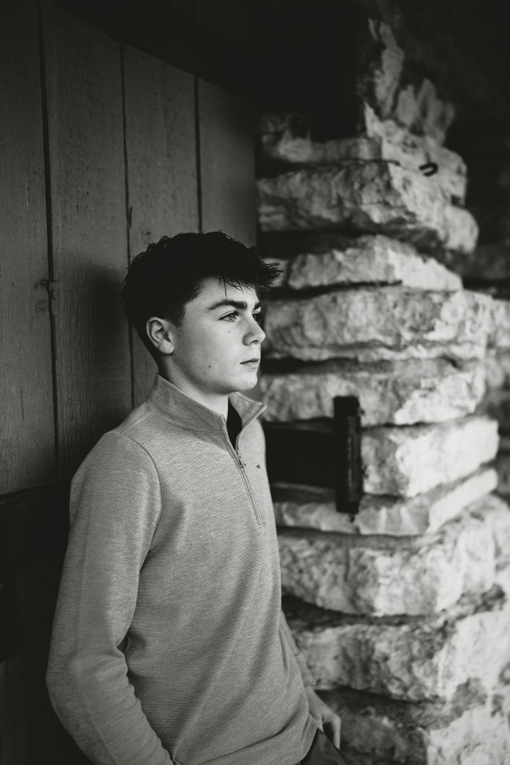 Black and white portrait of a young man in a quarter-zip pullover leaning against a rustic stone wall and wood paneling, gazing aside.