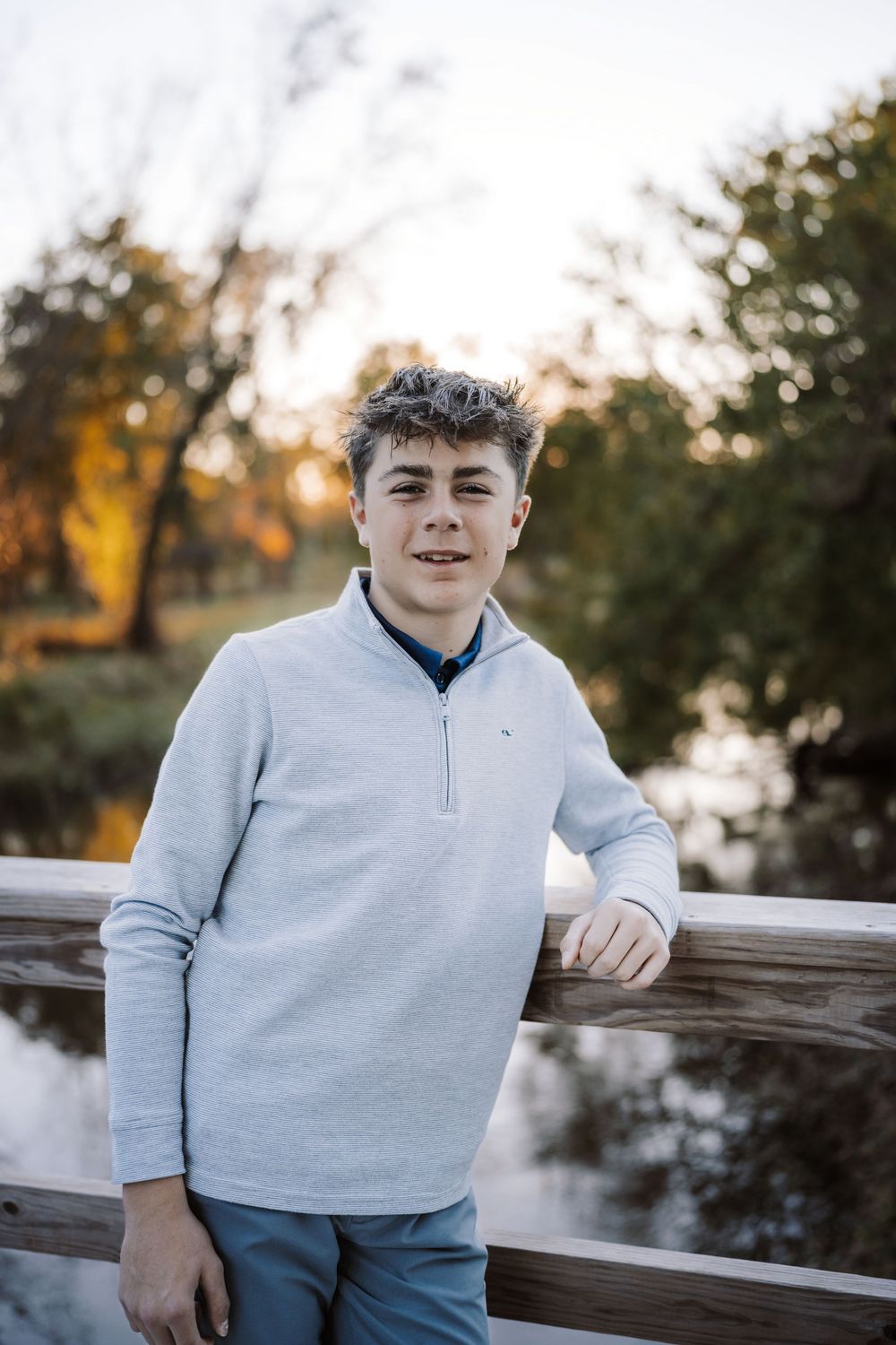 Smiling teenage boy in a light gray quarter-zip pullover leans on a wooden railing near a pond with golden autumn trees behind him.
