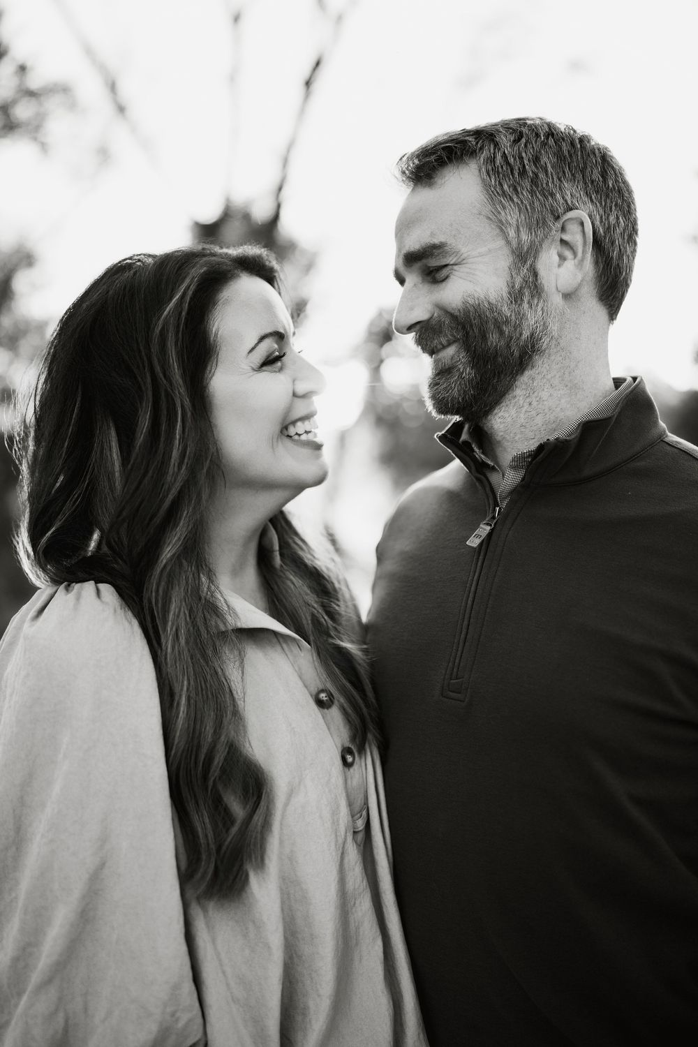 Black and white portrait of a smiling couple facing each other outdoors, the woman with long wavy hair and the bearded man in a zip pullover