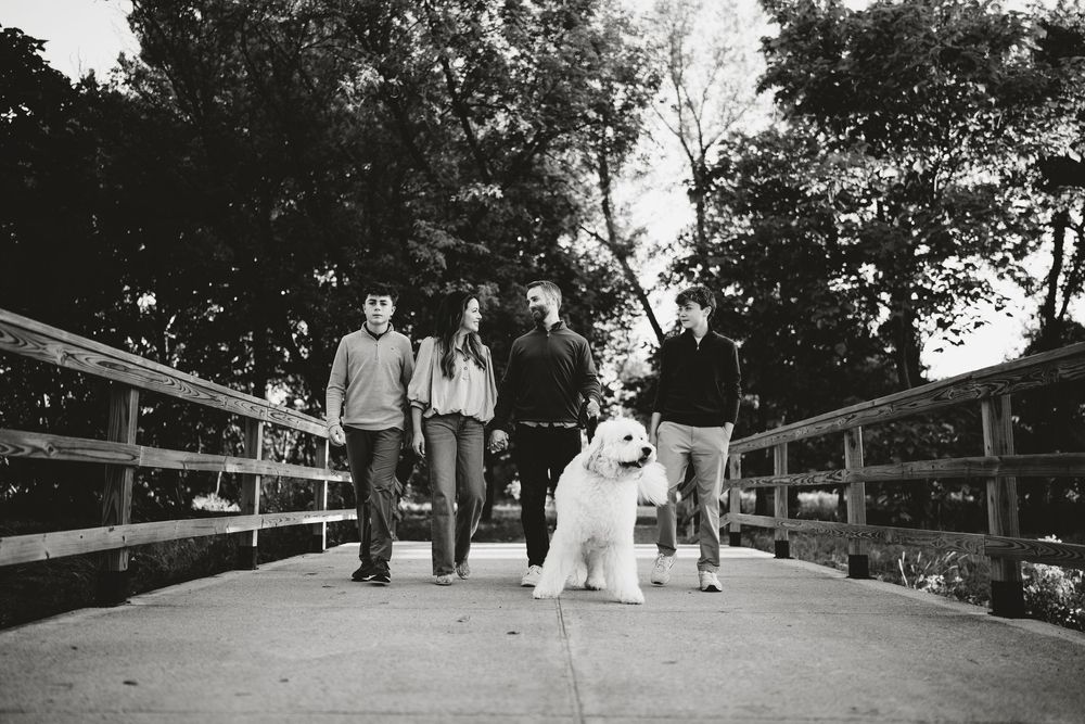 Black and white photo of a family of four walking with a large fluffy white dog across a wooden bridge surrounded by trees ion Oakbrook,IL