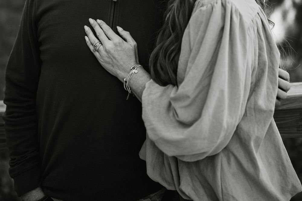 Close-up of a woman in a flowing blouse resting her hand with a bracelet and rings on a man's dark sweater in an intimate embrace.