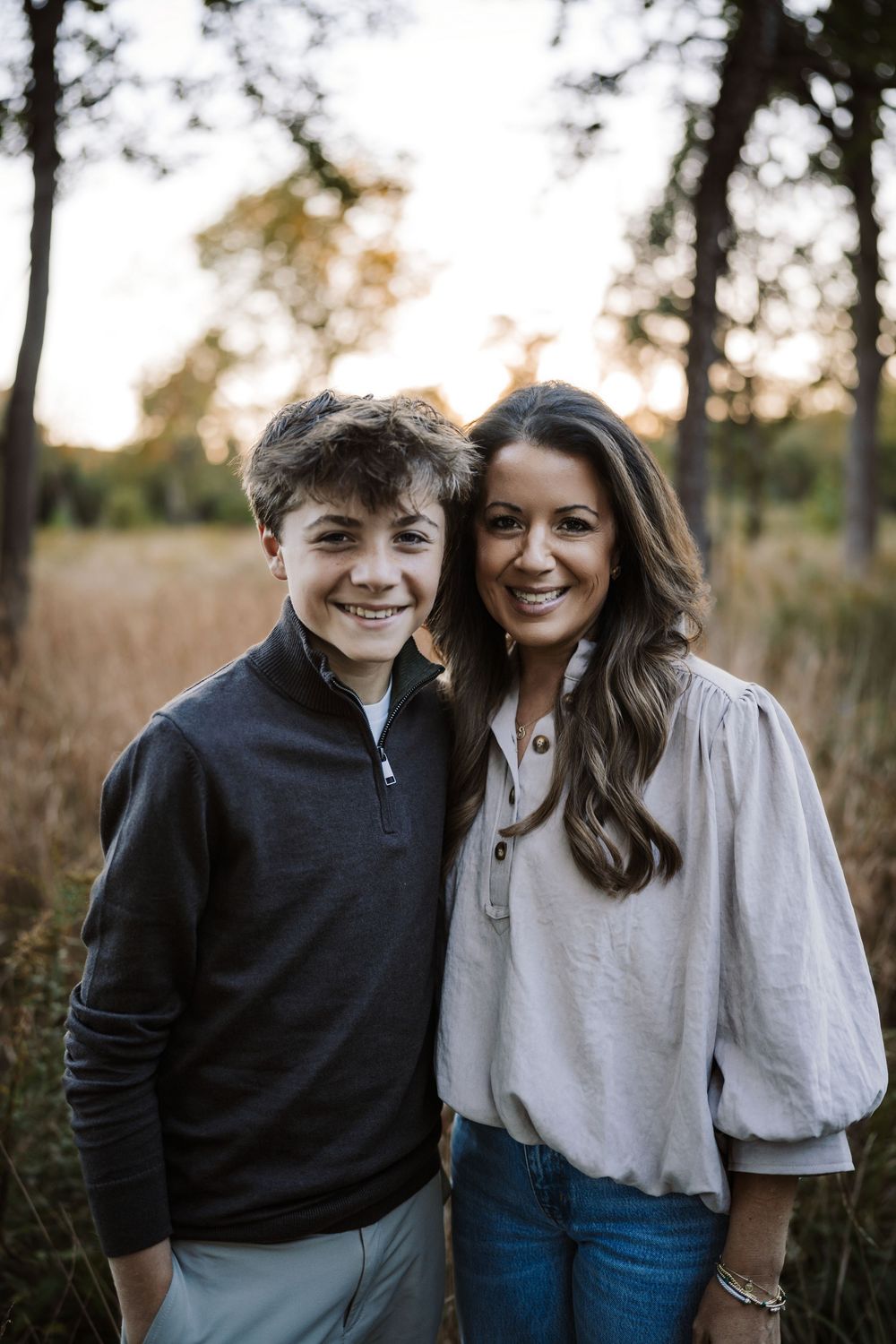 A smiling boy in a gray quarter-zip pullover poses beside a smiling woman in a blouse and jeans in a golden field at sunset.