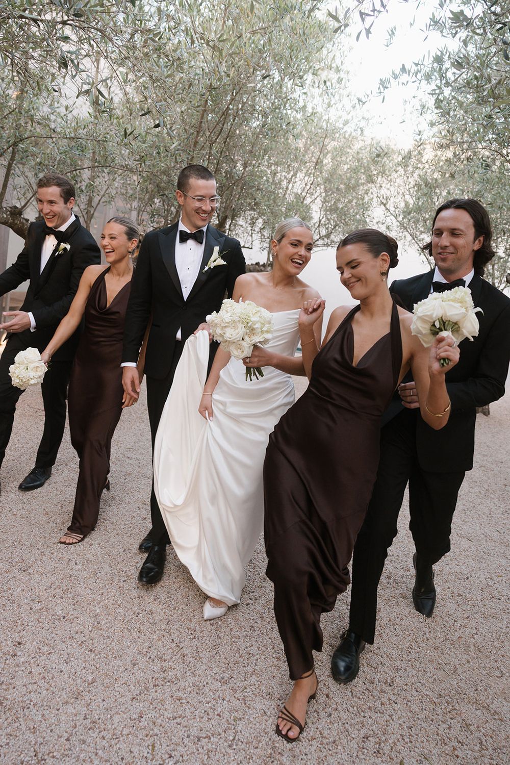Wedding party walks outdoors in elegant attire, bride in white gown with bridesmaids in brown dresses and groomsmen in tuxedos.