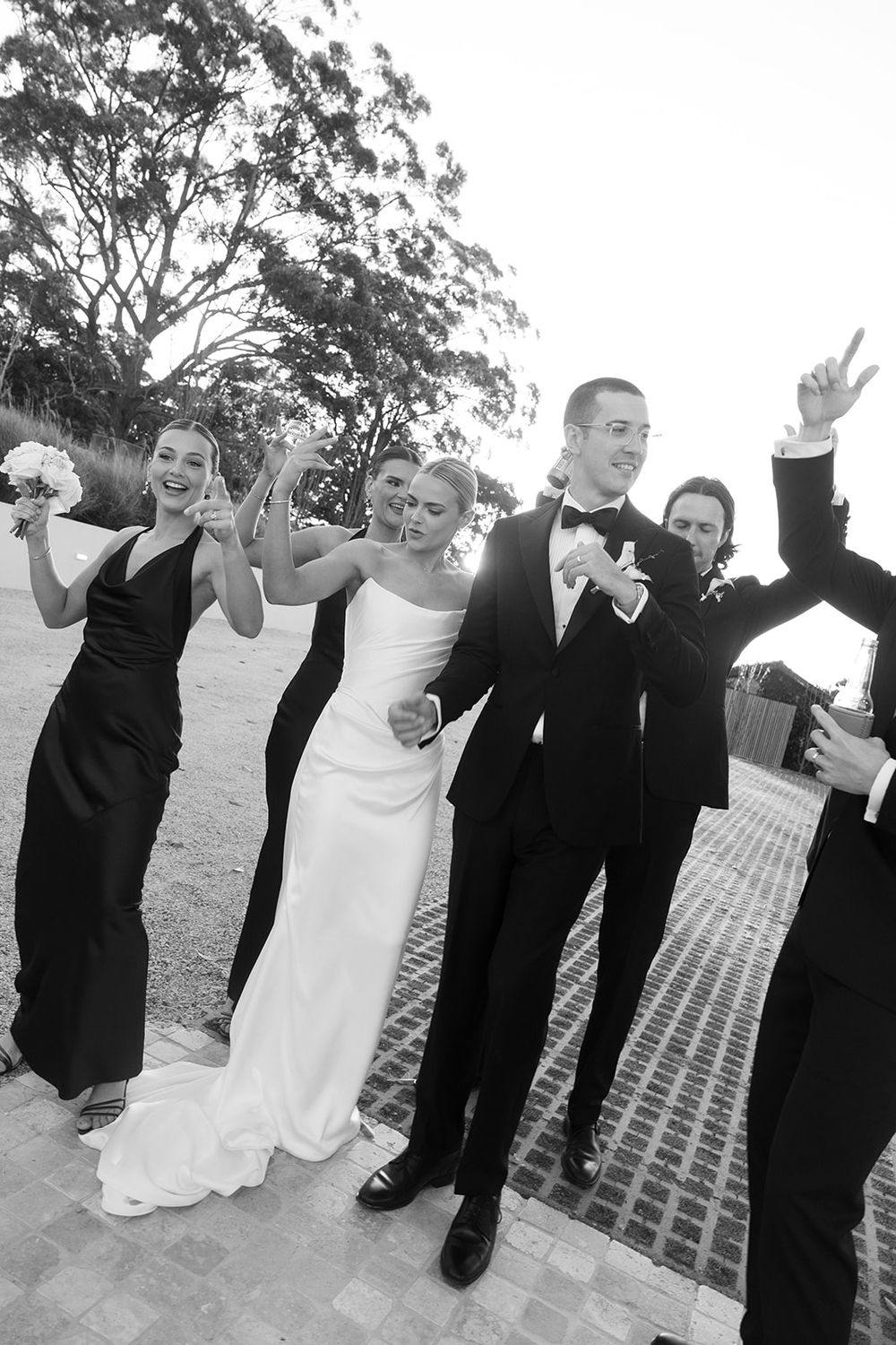 Black and white photo of a bride and groom walking with wedding party, bridesmaids in black gowns celebrating outdoors.