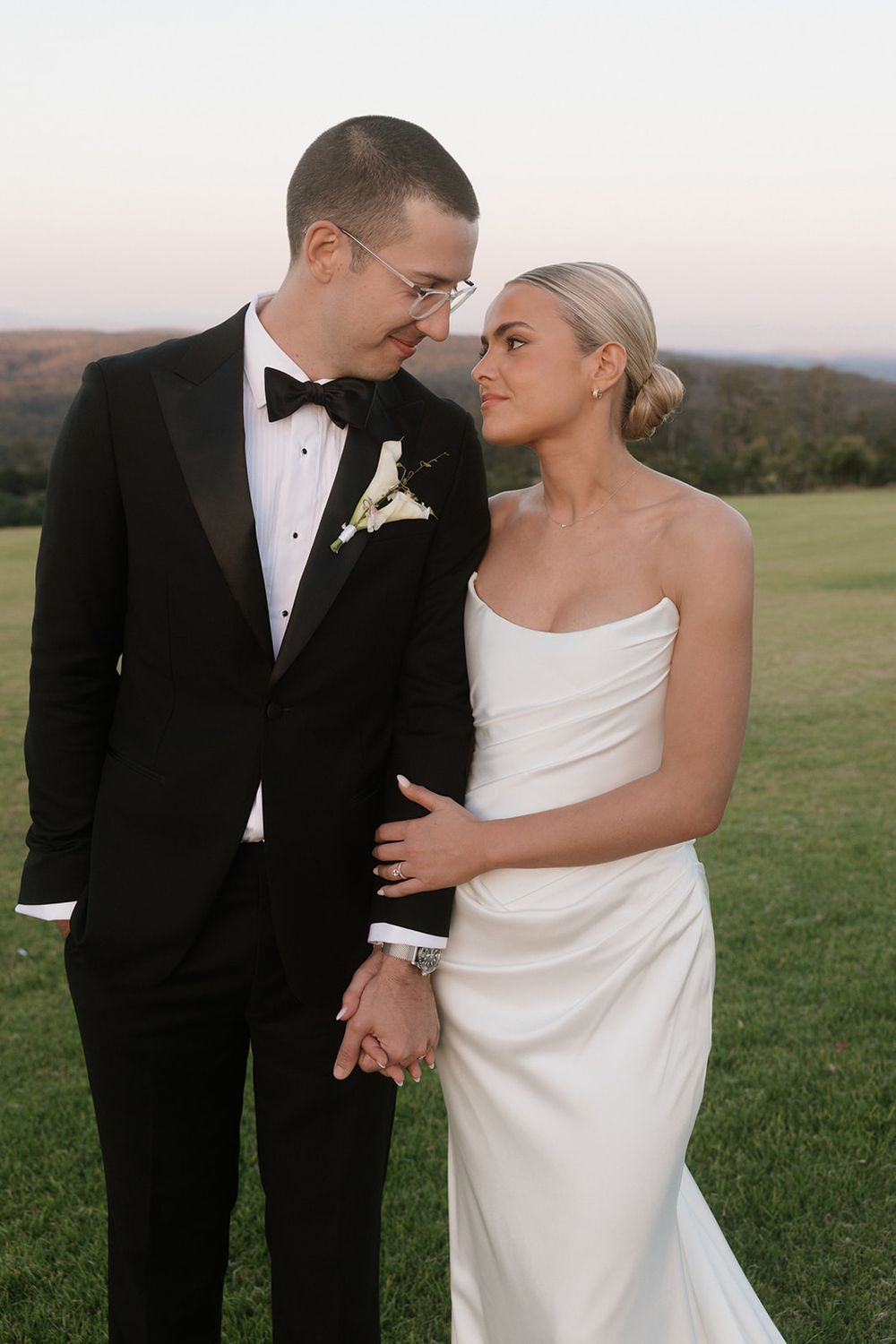 A couple in wedding attire share a romantic moment outdoors, groom in black tuxedo and bride in white strapless gown.