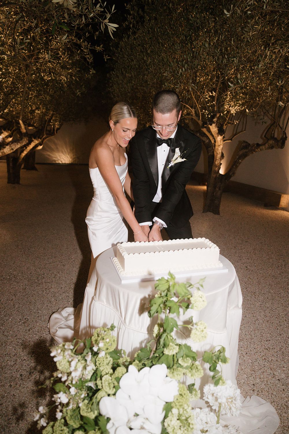 Bride and groom cutting white wedding cake outdoors at night, surrounded by olive trees and white floral arrangements.