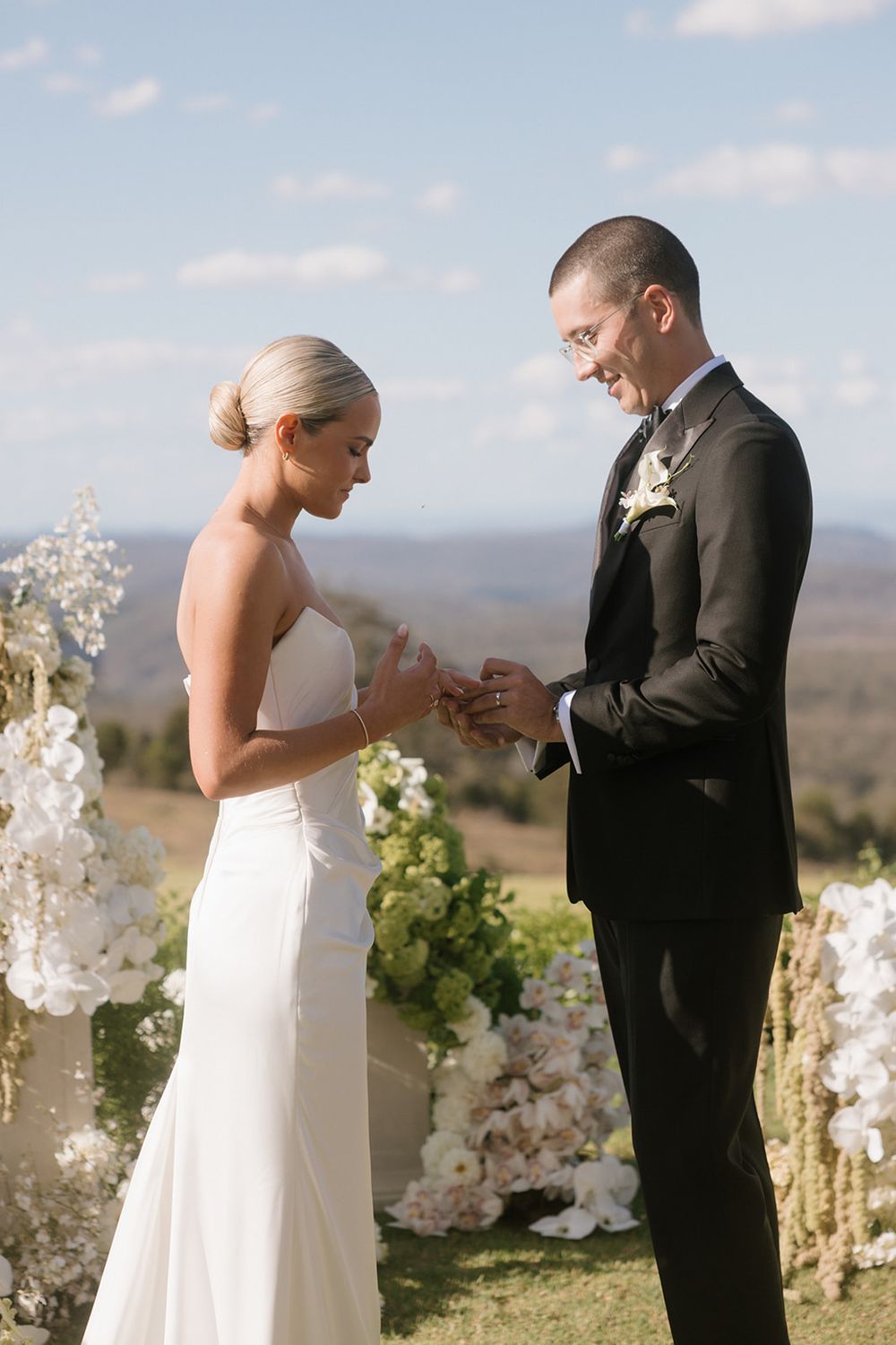 Bride places wedding ring on groom's finger during outdoor ceremony with floral arch and mountain views.