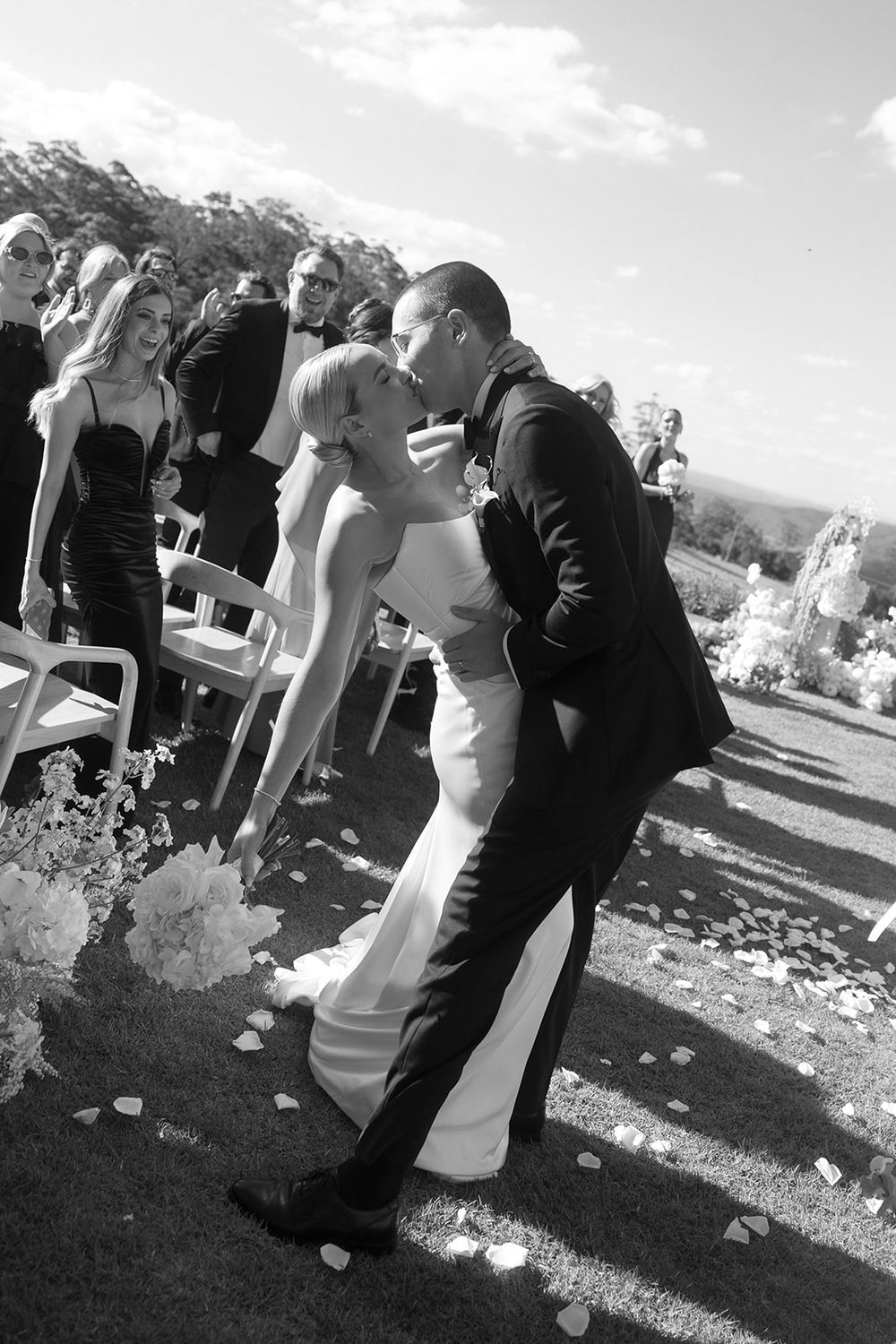 Black and white photo of bride and groom sharing a romantic kiss at outdoor wedding ceremony with guests watching.
